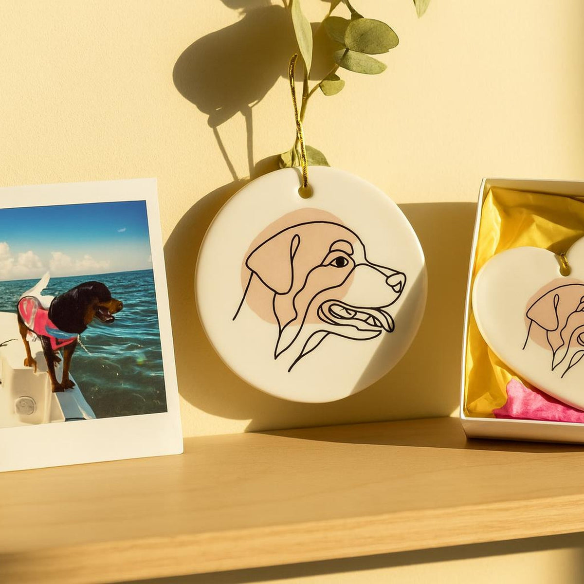A snapshot of a black-and-tan dog beside the round ceramic ornament printed with her one-line portrait, and a heart ornament in a white gift box