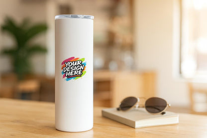 White 22oz skinny tumbler with a clear flip lid on a café counter, printed with the colourful paint-splash Your Design Here artwork